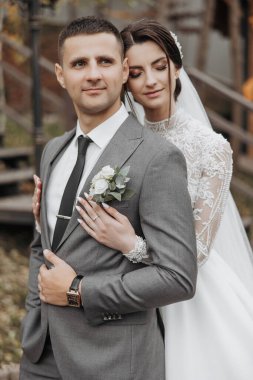 Düğün pozu veren bir erkek ve kadın. Adam takım elbise ve kravat takıyor ve kadın beyaz bir elbise giyiyor. İkisi de gülümsüyor ve mutlu görünüyorlar.