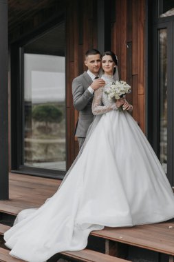 Gelin ve damat ahşap bir güvertede fotoğraf için poz veriyorlar. Gelin beyaz bir elbise giyiyor ve elinde bir buket çiçek tutuyor. Sahne mutlu ve kutlama dolu.