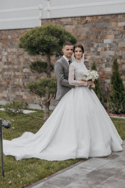 Bir gelin ve damat bir ağacın önünde fotoğraf için poz veriyorlar. Gelin bir buket tutuyor ve damat beyaz bir çiçek tutuyor. Sahne mutlu ve romantik.