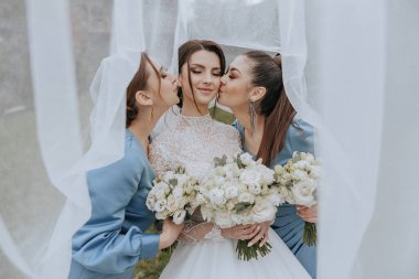 Üç kadın beyaz çiçekleri tutarken sarılıp öpüşüyorlar. Gelin mavi bir elbise giyiyor ve diğer iki kadın mavi elbiseler giyiyor. Sahne açık havada çekiliyor.