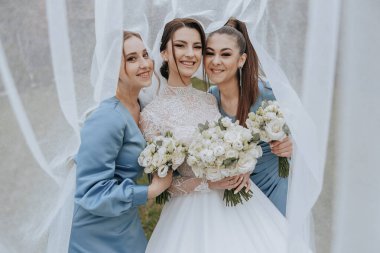 Üç kadın bir resim için poz veriyor, biri elinde bir buket çiçek tutuyor. Sahne neşeli ve kutlayıcı, çünkü kadınlar büyük ihtimalle bir düğüne ya da diğer özel etkinliklere katılıyorlar.