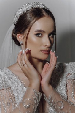 Kafasında taç ve boynunda kolye olan bir kadın. Beyaz bir elbise giyiyor ve elleri yüzünde.