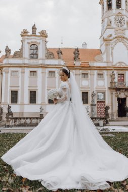 Uzun beyaz elbiseli bir gelin saat kulesi olan bir binanın önünde duruyor. Elinde bir buket çiçek tutuyor.