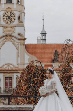 Beyaz bir elbise ve duvağı olan bir gelin saat kulesinin önünde duruyor. Elinde bir buket çiçek tutuyor.