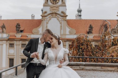 Gelin ve damat bir binanın balkonunda duruyorlar, gelin beyaz bir elbise giyiyor ve damat takım elbise giyiyor.