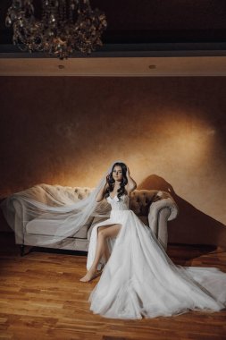 Gelinlikli bir kadın kanepede oturur. Kanepe beyaz ve çiçek desenli. Kadın peçe takıyor ve saçı topuz şeklinde. Oda loş bir şekilde aydınlatılmış, romantik bir ortam yaratıyor.