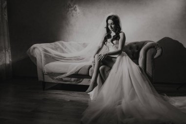 Gelinlikli bir kadın kanepede oturur. Kanepe beyaz ve siyah çerçeveli. Kadın bir taç takıyor ve kameraya bakıyor.