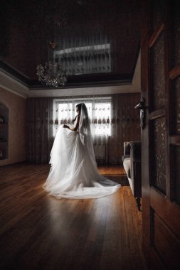 Gelinlikli bir kadın pencerenin önünde duruyor. Oda loş ve kadının elbisesi beyaz.