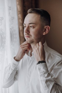 Bir adam resmi bir olaya hazırlanıyor, beyaz bir gömlek giyiyor ve kravatını düzeltiyor.