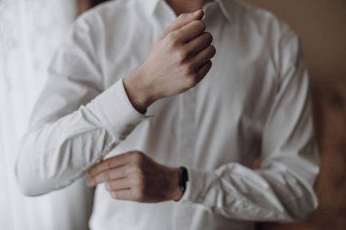 Bir adam beyaz gömlek giyiyor ve gömleğinin kollarını düzeltiyor. Formalite ve detaylara dikkat etme kavramı, çünkü adam uygun bir şekilde giyinmek için zaman harcıyor.