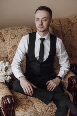 Siyah yelek ve beyaz gömlekli bir adam kanepede oturuyor. Siyah bir kravat takıyor ve bileğinde bir saat var. Önüne bir buket çiçek konur.