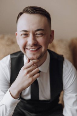 Bir adam gülümsüyor ve siyah yelek ve beyaz gömlek giyiyor. Kanepede oturuyor.