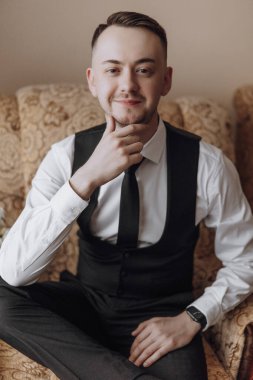 Siyah yelek ve beyaz gömlekli bir adam kanepede oturuyor. O bir kravat takıyor ve yüzünde bir gülümseme var.