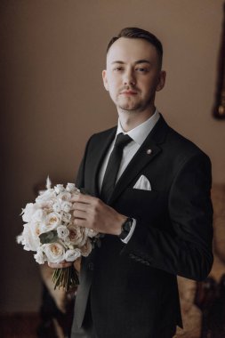 Elinde bir buket çiçek tutan takım elbiseli bir adam. Adam siyah bir takım elbise ve beyaz bir gömlek giyiyor. Buketi sağ elinde tutuyor.