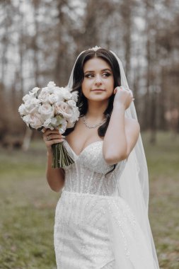 Beyaz bir gelinlik giyen ve elinde bir buket çiçek tutan bir kadın. Arka planda ağaçlarla bir tarlada duruyor.