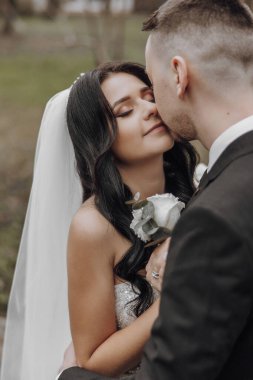 Bir gelin ve damat birbirlerine sarılıp öpüşüyorlar. Gelin elinde bir çiçek tutuyor.