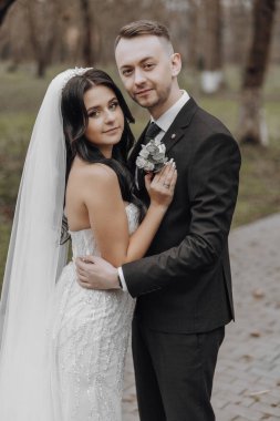 Bir gelin ve damat parkta fotoğraf için poz veriyorlar. Gelin beyaz bir elbise giyiyor ve damat bir takım elbise giyiyor. İkisi de gülümsüyor ve mutlu görünüyorlar.