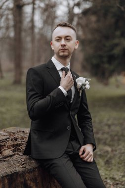 Takım elbiseli ve kravatlı bir adam ormanda bir kütüğe oturur. Yakasına beyaz bir çiçek takmış.