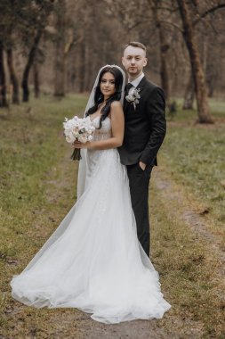 Bir gelin ve damat ormanda fotoğraf çektirmek için poz veriyorlar. Gelin beyaz bir elbise giyiyor ve elinde bir buket çiçek tutuyor. Sahne romantik ve samimi.