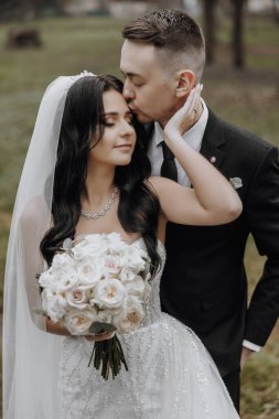 Bir gelin ve damat birbirlerinin boyunlarını öpüyor. Gelin elinde bir buket çiçek tutuyor.