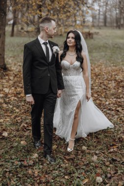 Gelin ve damat ormanda yürüyor, gelin beyaz bir elbise ve damat siyah bir takım elbise giyiyor.