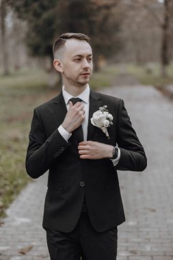 Takım elbiseli bir adam yakasına beyaz bir çiçek takıyor. Tuğla bir geçitte duruyor.