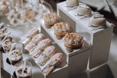 Pasta, tart ve diğer tatlı ikramlardan oluşan bir masa. Tatlılar beyaz bir masada düzenlenir ve küçük bardaklarda sunulur. Masa çeşitli tatlılarla süslenmiştir.