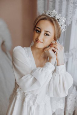 Bir kadın düğün için hazırlanıyor ve tacını takıyor. Beyaz bir elbise giyiyor ve bir pencerenin önünde duruyor.