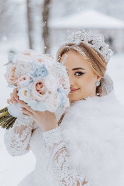 Bir gelin, elinde bir buket çiçek ve taç olan bir resim için poz veriyor. Beyaz bir kürk giyiyor ve yüzü kısmen çiçeklerle kaplı. Zarafet ve güzellik kavramı