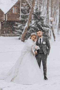 Bir gelin ve damat karda fotoğraf için poz veriyorlar. Damat takım elbise giyerken gelin beyaz bir elbise ve kürk şalı giyiyor. Bir evin önünde duruyorlar.