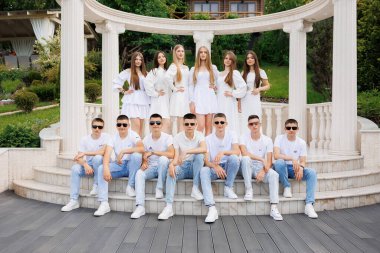 Bir grup genç beyaz bir kemer yolunun önünde fotoğraf için poz veriyor. Sahne neşeli ve eğlenceli, grup beyazlar içinde kameraya gülümsüyor.