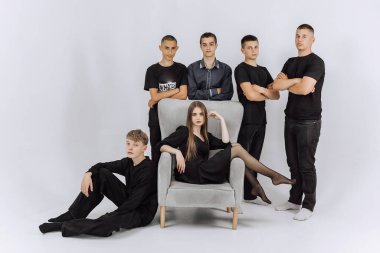 Bir grup genç erkek ve kadın bir sandalyenin önünde fotoğraf için poz veriyorlar. Sandalye grubun ortasında, etrafında insanlar dikilirken konumlandırılmıştır. Olay yeri sıradan ve dostane.