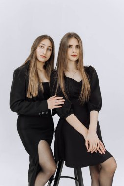 Siyah elbiseli iki genç kadın fotoğraf için poz veriyor. Aynı kıyafetleri giyiyorlar ve bir taburede oturuyorlar. Sahne zarif ve sofistike.