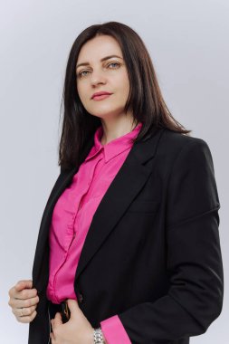 Siyah takım elbiseli ve pembe gömlekli bir kadın fotoğraf için poz veriyor. Bileğinde bir saat ve pembe bir gömlek var.