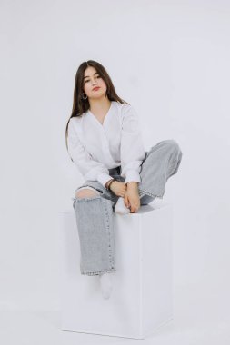 Bir kadın yırtık kotlu beyaz bir kutunun üzerinde oturuyor. Üzerinde beyaz bir gömlek var ve bilezik takıyor.