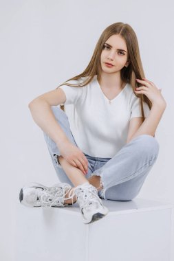 Bir kadın bacak bacak üstüne atmış beyaz bir kutunun üzerinde oturuyor. Beyaz bir gömlek ve kot pantolon giyiyor.