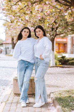 İki kadın bir ağacın önünde poz veriyor. Kot pantolon ve beyaz gömlek giyiyorlar.