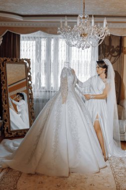Bir gelin düğünü için hazırlanıyor ve bir aynanın önünde duruyor. Uzun beyaz bir elbise giyiyor, üstünde tren ve duvak var.
