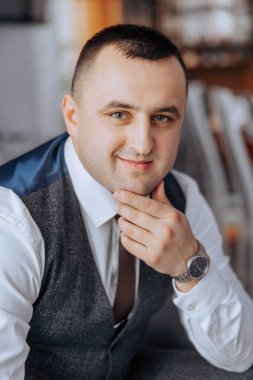 Mavi yelek ve beyaz gömlek giyen bir adam gülümsüyor ve kameraya bakıyor. Bileğine saat takmış.