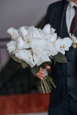 Bir adam elinde bir buket beyaz gül tutuyor. Adam takım elbise ve kravat takıyor.