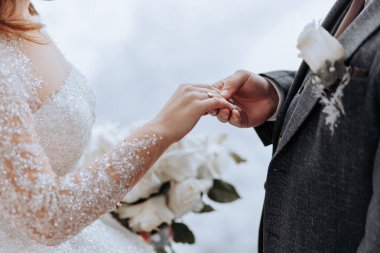 Bir gelin ve damat evleniyor ve gelin elinde bir buket beyaz çiçek tutuyor. Damat gelinin parmağına yüzük takmak üzere.