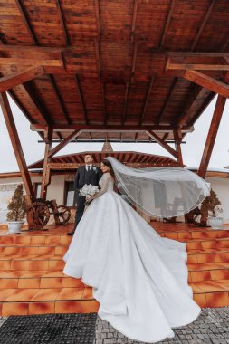 Bir gelin ve damat binanın önündeki merdivenlerde dikiliyorlar. Gelin uzun beyaz bir elbise giyiyor ve damat bir takım elbise giyiyor. Sahne romantik ve mutlu.