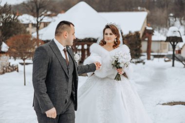Gelin ve damat karda yürüyorlar, gelin kürk manto giyiyor ve bir buket tutuyor.