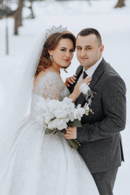 Bir gelin ve damat karda fotoğraf için poz veriyorlar. Damat takım elbise giyerken gelin beyaz bir elbise ve taç giyiyor. Bir buket çiçek ve çiçek tutuyorlar.