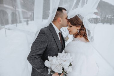 Bir gelin ve damat karda öpüşüyor. Gelin beyaz bir kürk giyiyor ve damat da takım elbise giyiyor.