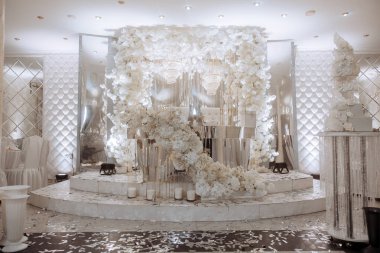 Beyaz bir çiçek kemeri ve beyaz bir masası olan büyük bir odada düğün resepsiyonu düzenleniyor. Oda beyaz çiçekler ve mumlarla süslenmiş. Romantik ve zarif bir ortam yaratıyor.