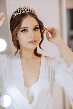 Bir kadın taç ve beyaz bir bornoz giyiyor. Aynada kendine bakıyor.