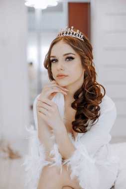 Taçlı ve beyaz cüppeli bir kadın yatağa oturur. Kendinden emin ve dengeli görünüyor.