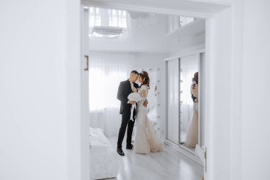 Gelin ve damat beyaz bir kapının önünde, gelin bir buket tutuyor. Sahne romantik ve samimi, çünkü çift birbirini kucaklıyor ve gözlerinin içine bakıyor.