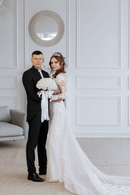 Bir gelin ve damat beyaz bir odada fotoğraf için poz veriyorlar. Gelin uzun beyaz bir elbise giyiyor ve damat siyah bir takım elbise giyiyor. Sırasıyla bir buket ve kravat tutuyorlar.